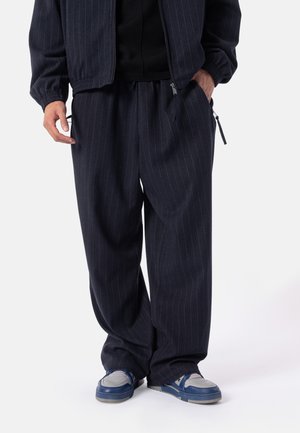 Persoon draagt losse donkere broek met krijtstreep, bijpassend jasje, zwart shirt en blauw-witte sneakers, staat met handen in de zakken.