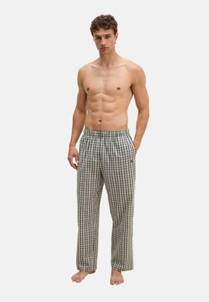 Junger Mann steht barfuß, trägt lockere karierte Pyjamahose mit den Händen in den Taschen vor einem schlichten hellen Hintergrund.
