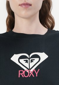 Black cotton t-shirt featuring a large white heart graphic and the text "ROXY" in pink. Round neckline and short sleeves.