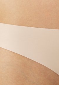 Beige sömlös stringtrosa med en slät textur. Har en bred midjeband och en minimalistisk design som visar en subtil lyster.