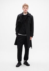 Schwarzer Trenchcoat mit doppelreihigen Knöpfen, getragen über einem schwarzen Hemd und Jogginghosen. Schwarze Sneakers vervollständigen das Outfit. Einfaches, minimalistisches Design.