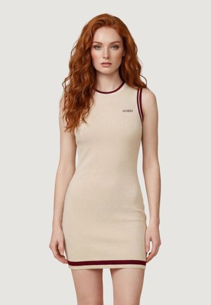 Donna dai capelli rossi che indossa un vestito senza maniche beige con rifiniture color bordeaux e il logo "GUESS", in piedi davanti a uno sfondo chiaro e semplice.