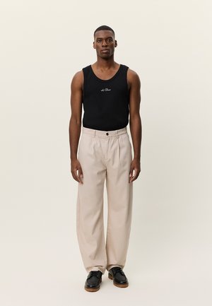 Άνδρας που στέκεται με το βλέμμα προς τα εμπρός φορώντας μαύρο αμάνικο τοπ, μπεζ παντελόνι με φαρδύ πόδι και μαύρα παπούτσια σε απλό φόντο.