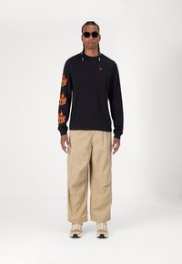 Schwarzes Langarmshirt mit orangefarbenen Flammenmotiven auf den Ärmeln, kombiniert mit beigen, weit geschnittenen Cargo-Hosen und beigen Sneakers.