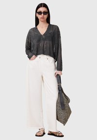 Schwarzes halbtransparentes V-Ausschnitt-Oberteil, weiße weite Hosen mit fransigem Saum, metallische Sandalen und eine gemusterte Tasche mit einem schwarzen Tragegurt.