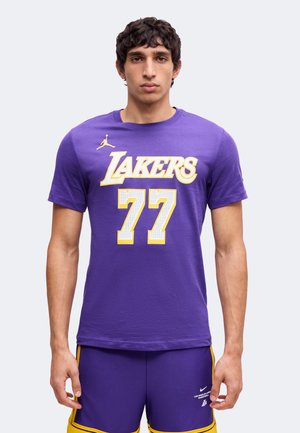 Jeune homme portant un t-shirt violet des Los Angeles Lakers avec le numéro 77 et un short assorti violet et jaune, debout sur un fond clair.