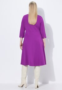 Robe violette avec des manches trois quarts, une jupe évasée et un dos épuré. Associée à des bottes blanches montantes.