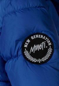 Blå puffjacka av tyg med kviltad struktur, prydd med en cirkulär svart broderad patch som läser "NYA GENERATIONEN" och "Mnoti."