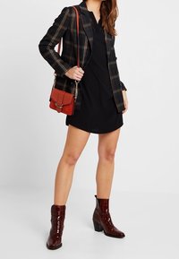 Robe noire associée à un blazer à carreaux. Les accessoires comprennent un petit sac rouge et des bottines rouges brillantes avec un motif textur é façon crocodile.