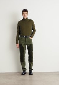 Olive green turtleneck sweater, paired with olive green textured pants featuring darker patches. Worn with black shoes, model stands against a neutral backdrop.