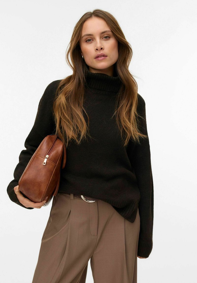 Ciemnozielony dzianinowy sweter z wysokim kołnierzem, zestawiony z beżowymi plisowanymi spodniami. Modelka trzyma brązową skórzaną torbę z zamkiem błyskawicznym.