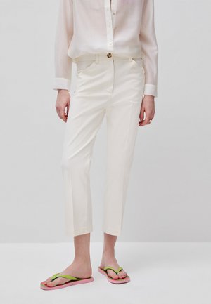 Person trägt hellbeige gekürzte Hose, durchscheinendes hellbeige Hemd mit Knöpfen und pinke Flip-Flops mit grünen Riemen vor einem einfarbigen Hintergrund.