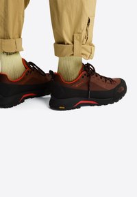 Persona che indossa pantaloni beige risvoltati, calzini verde chiaro e scarpe da trekking marrone-nero con dettagli rossi su sfondo bianco.