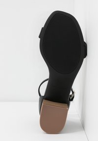 Vue de dessous d’une sandale noire avec une semelle texturée et un petit talon bloc marron appuyée contre des murs blancs.