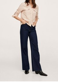 Chemise rose clair boutonnée avec deux poches poitrine, associée à un jean large bleu foncé et des bottines noires à bout pointu.