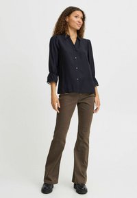 Camicia nera con bottoni e maniche a sbuffo tre quarti, in tessuto leggero e con un'elegante dettaglio ai polsini, abbinata a pantaloni marroni a campana e stivali neri.