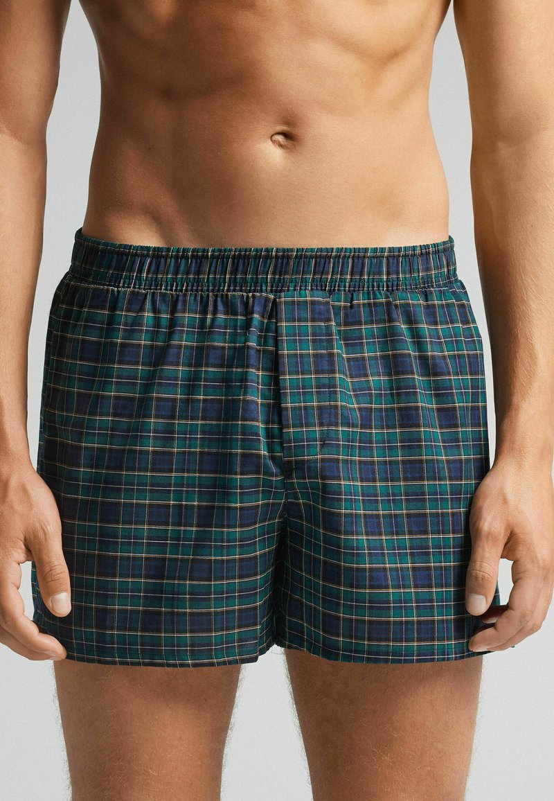 Pánské trenýrky vyrobené z bavlny, s tmavě modrým a zeleným kostkovaným vzorem a žlutými akcenty, s elastickým pasem a uvolněným střihem.