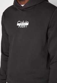 Felpa grigia scura con tasca frontale, caratterizzata da un logo bianco di Calvin Klein. Realizzata in misto cotone, ha una texture morbida e una vestibilità standard.