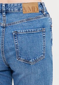 Jeans iz denima v svetlo modri barvi z teksturiranim zaključkom, ki vsebuje značilno svetlo rjavo usnjeno oznako z napisom "VMD" in en sam zadnji žep.