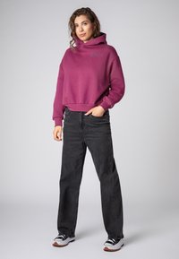 Bordeaux hoodie met een voorkeurszak, losse pasvorm, en geribde onderrand, gecombineerd met zwarte rechtse jeans en zwart-witte sneakers.