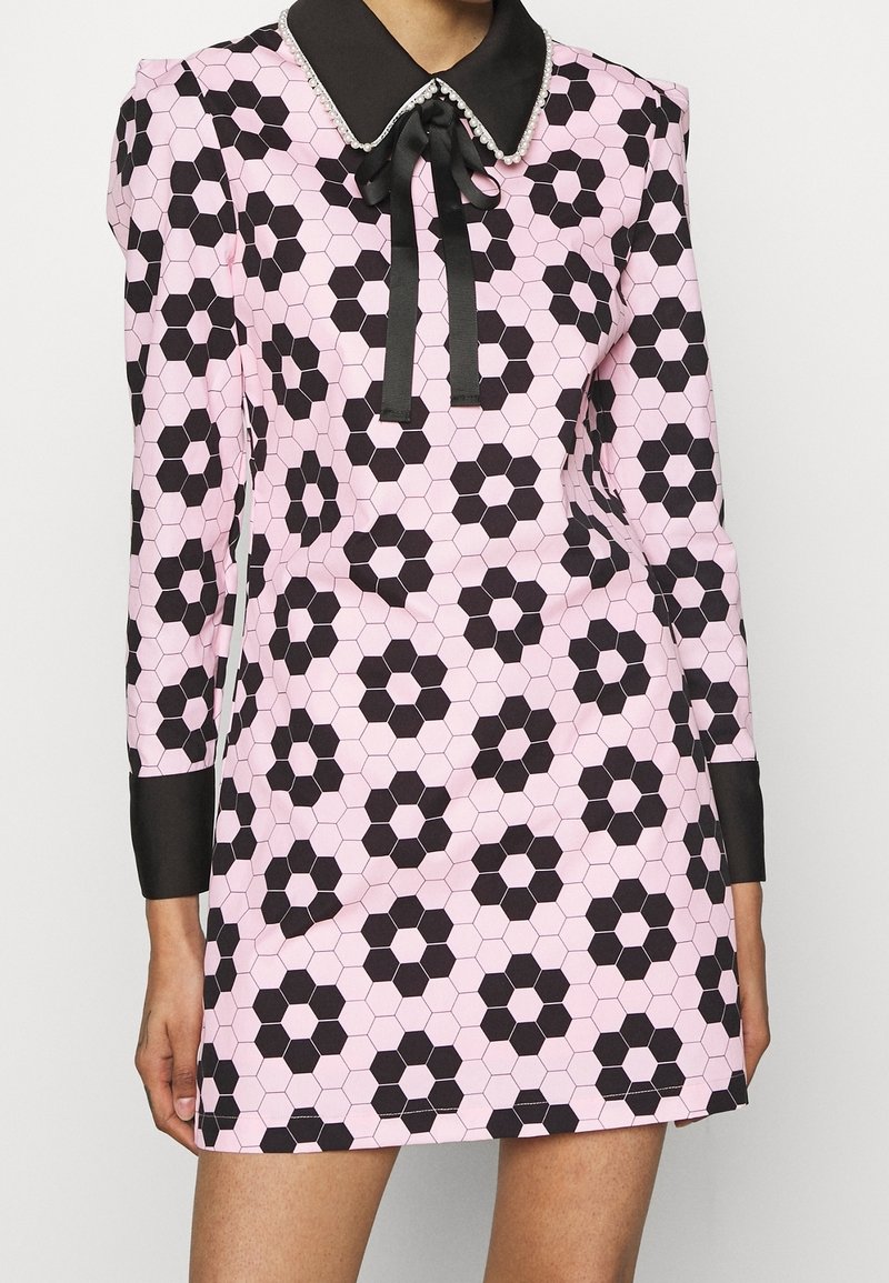 Rosa klänning med svart hexagonal mönster, långa ärmar, svart krage och nackslips. Har dekorativa detaljer på kragen.