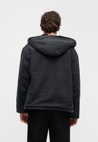 Μαύρο μπουφάν με κουκούλα και υφή, με άνετη εφαρμογή και ορατές ραφές. Η οπίσθια όψη παρουσιάζει το σχέδιο της κουκούλας και του τελειώματος.