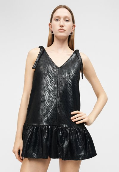 Kobieta ubrana w czarną, bezrękawnikową sukienkę z imitacji skóry węża, z wiązaniami na ramionach i falbaną u dołu, stojąca na jednolitym tle.