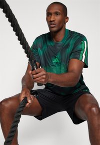 Πράσινη αθλητική μπλούζα από διχτυωτό υλικό με υφή, με λευκές λεπτομέρειες, συνδυασμένη με μαύρες βερμούδες. Άτομο κρατάει ένα χοντρό, μαύρο σχοινί.