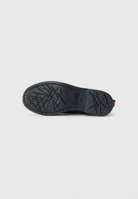 Semelle de chaussure noire avec motif de crampons croisés, bout arrondi et logo de la marque embossé près du talon, sur un fond clair uni.