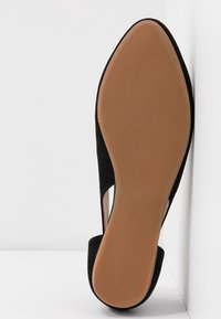 Chaussure plate noire à bout pointu, semelle couleur tan, présentée sur le côté contre un fond blanc d'angle.