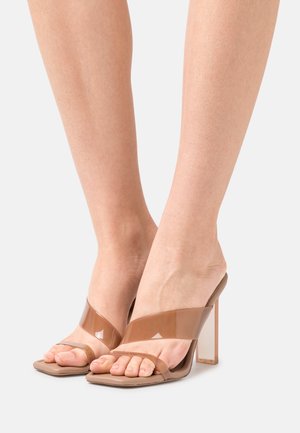 Gennemsigtige hælede sandaler med en beige sål, der har krydskrydsede remme og en blokhæle. Glat tekstur og moderne design.