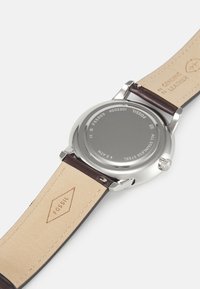 Montre en acier inoxydable argenté avec un bracelet en cuir marron. L'arrière a une surface lisse avec des détails gravés ; elle possède un boîtier rond et une couronne.