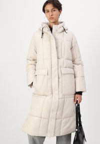 Lys beige polstret kåpe med hette, frontlommer og trykknapper, med et quiltet design og en avslappet passform.