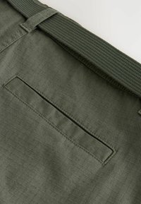 Gros plan sur une ceinture en tissu kaki avec une ceinture texturée et une poche passepoilée horizontale cousue sur le tissu.