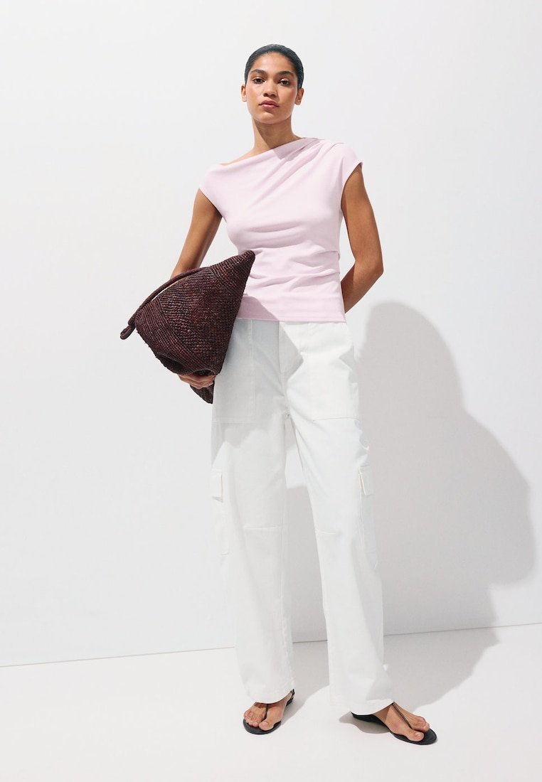 Femme debout contre un mur blanc portant un haut rose clair sans manches, un pantalon blanc à jambes larges, des sandales noires, tenant un grand sac tressé marron foncé.