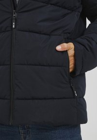 Granatowa puchowa kurtka z zamkiem z przodu, kieszeniami bocznymi i przeszytymi poziomymi liniami. Materiał wydaje się gładki i ocieplony.