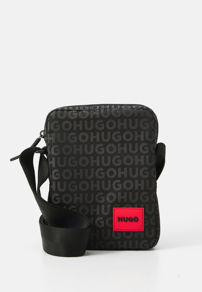 Small black crossbody bag with repeated "HUGO" text pattern and red square logo patch, featuring an adjustable black strap.