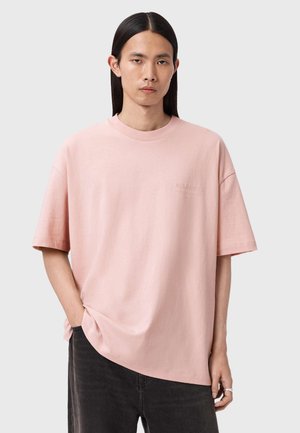 Ung person med langt mørkt hår iført en løs lyserød T-shirt og sorte jeans, stående med den ene hånd i lommen mod en ensfarvet baggrund.