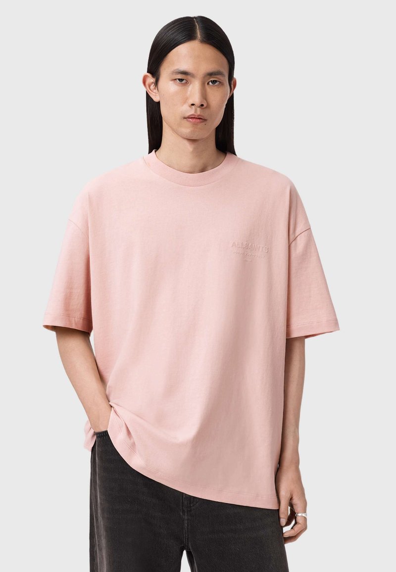 Junge Person mit langen dunklen Haaren, die ein locker sitzendes pinkfarbenes T-Shirt und schwarze Jeans trägt, mit einer Hand in der Tasche vor einem schlichten Hintergrund stehend.