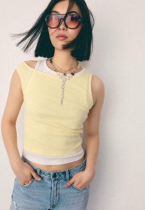 Frau mit schwarzem Bob-Haarschnitt, die übergroße rosa Sonnenbrillen trägt, geschichtete gelbe und weiße ärmellose Oberteile, blaue Jeans und eine Anhänger-Halskette.