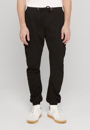 Man wearing black cargo jogger pants with drawstring waist and white sneakers standing against plain background.