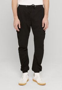 Bărbat purtând pantaloni cargo jogger negri cu talie cu șnur și adidași albi, stând în fața unui fundal simplu.