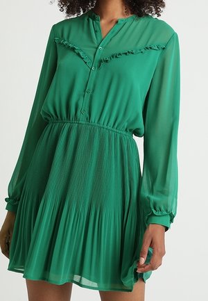 Vrouw draagt een groene jurk met lange mouwen, knopen, ruches op de borst en een geplooide rok, staand voor een effen achtergrond.