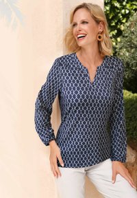 Marineblauwe blouse met een geometrisch wit patroon, lange mouwen, ronde halslijn en textuurstof. Gecombineerd met lichtgekleurde broeken.