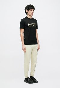 Juodas medvilninis marškinėlis su auksiniu „EA7 Emporio Armani“ logotipu, derinamas su kreminėmis sportinėmis kelnėmis ir juodais sportbačiais, vilkinčiu vyru modeliu.