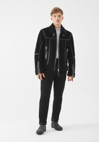 Giacca in suede nera con colletto in shearling, tasche con zip, cuciture a contrasto, indossata sopra una camicia grigia e abbinata a jeans neri e stivali grigi.