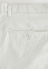 Poche arrière d'un pantalon beige clair avec une fermeture à un seul bouton et une boucle de ceinture au-dessus sur un fond uni.