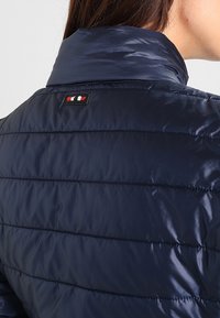 Mörkblå pufferjacka med hög krage, quiltat mönster och logotyp med röda, vita och blå accenter på baksidan.