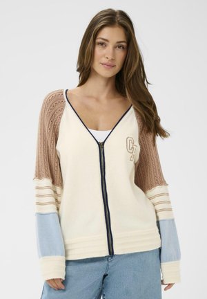 Lukuga kardigan, mille kere on kreemikas, tekstuursete beežide ja siniste varrukatega ning tumesinise äärega. Rinnaosas on „CR" logo.