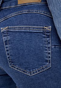 Jeans de denim azul oscuro con un bolsillo trasero con costuras en contraste y una etiqueta de cuero arriba. La textura parece suave con un leve desvanecimiento índigo.